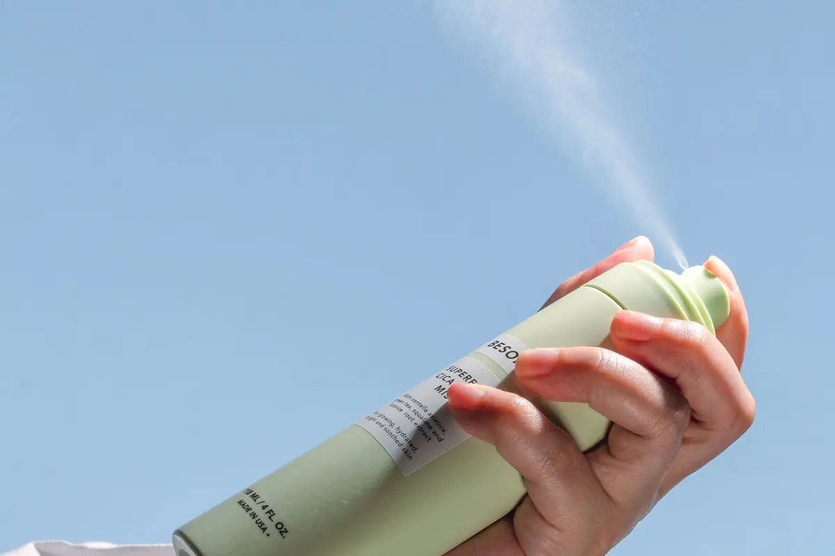 A hand holds up an aerosol can spraying a plume against a blue sky