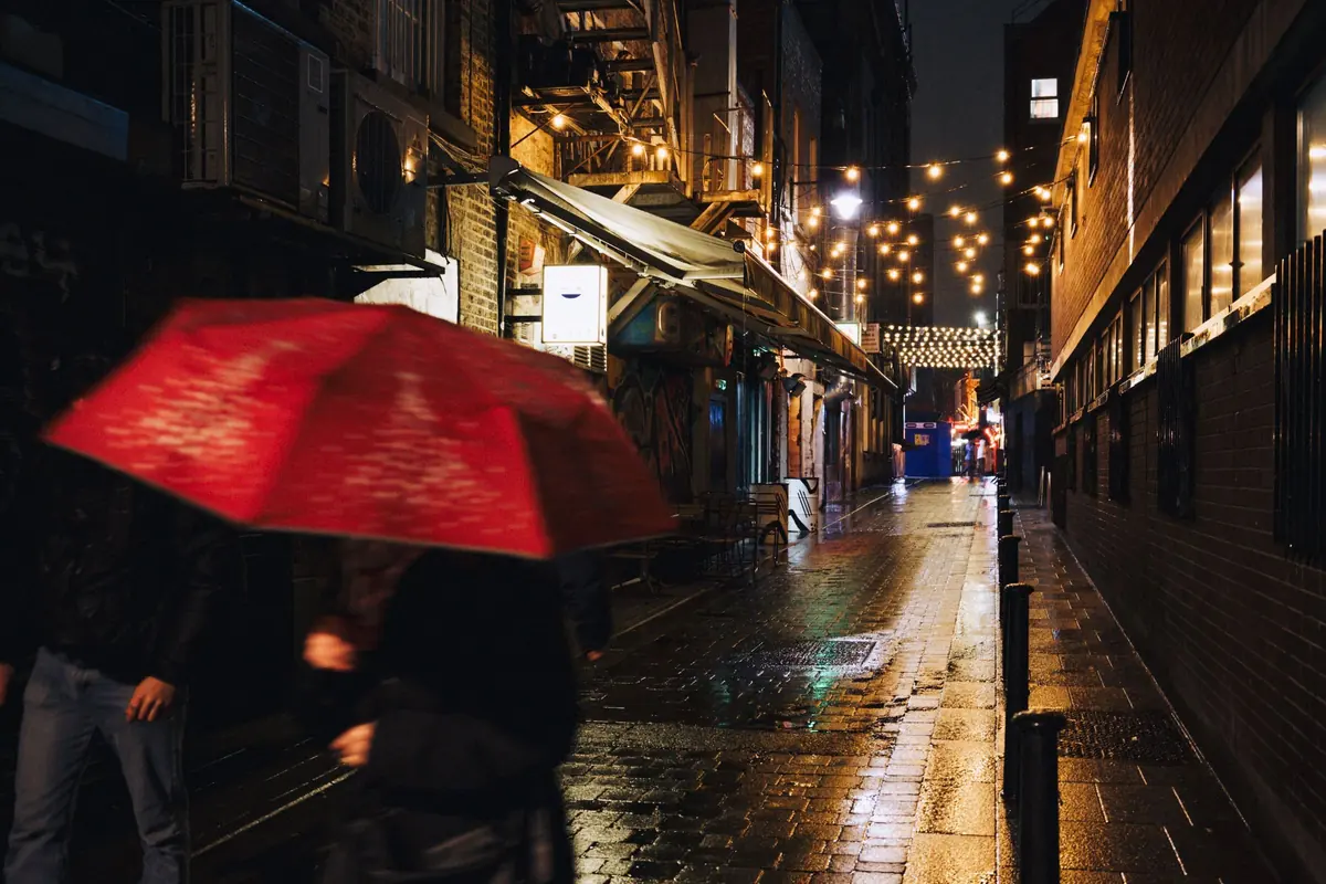 A person with a red umbrella walks down a wet, lamp-lit street at night