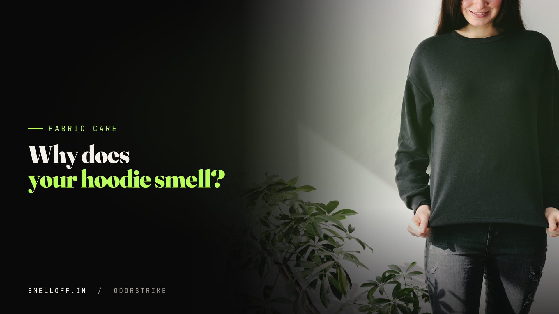 A woman in a plain black sweatshirt stands against a white wall beside a houseplant